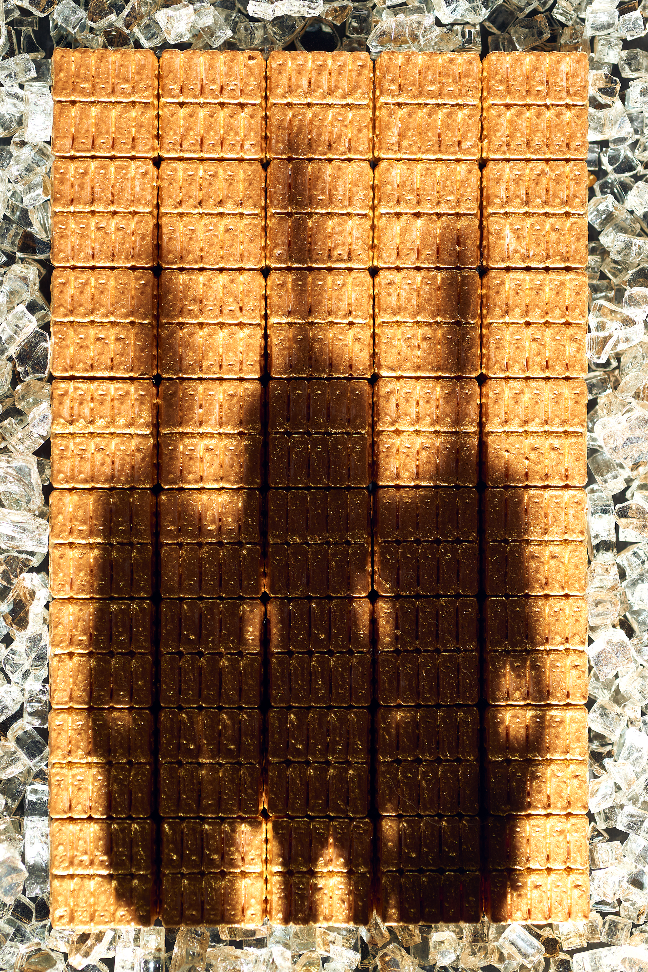 A studio photograph with what appears to be piles of gold bars that fill the majority of the frame and are framed perfectly symmetrical. Framing the gold bars are crushed diamonds or glass. The shadows of three men are cast across the gold bars, also in a symmetrical arrangement.